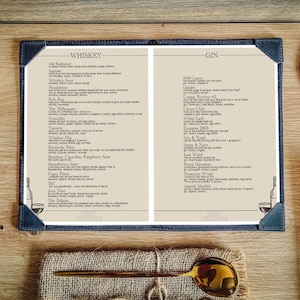 May include: A drinks menu with whiskey and gin cocktails, bound in dark blue leather. The menu is open, showing drink recipes. A gold spoon and a coaster are on a wooden table.