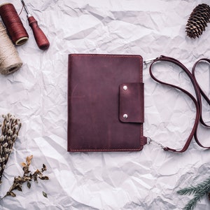 May include: A burgundy leather portfolio with a strap and a small pocket on the front. The portfolio is lying on a white crumpled paper background.