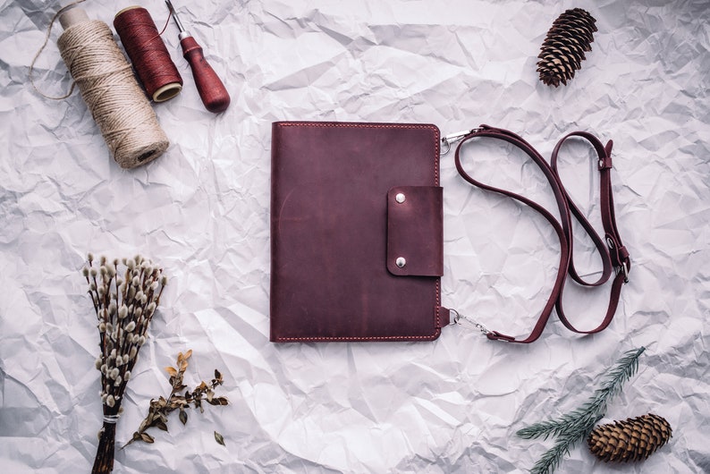 May include: A burgundy leather portfolio with a strap and a small pocket on the front. The portfolio is lying on a white crumpled paper background.