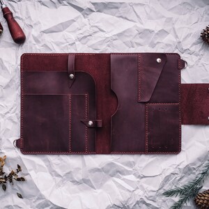 May include: A burgundy leather portfolio with multiple pockets and compartments, open and lying flat on a crumpled white paper background.