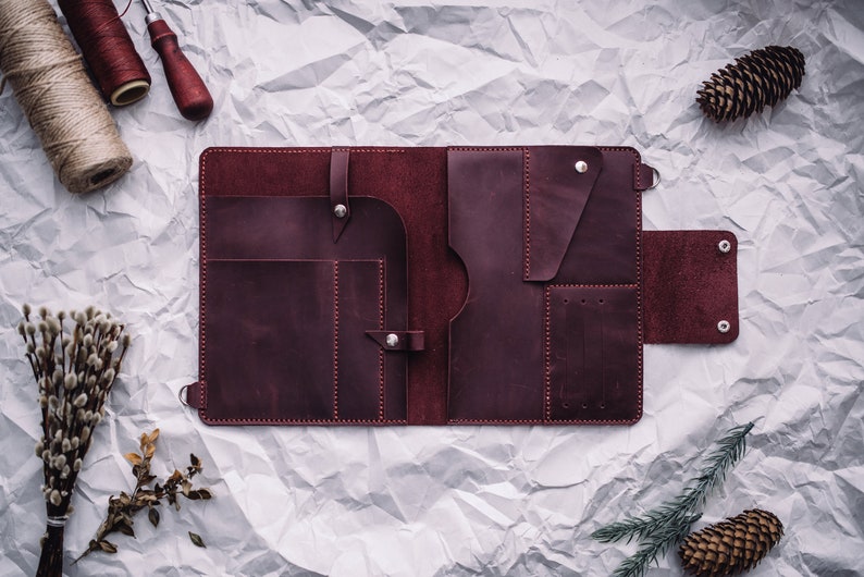 May include: A burgundy leather portfolio with multiple pockets and compartments, open and lying flat on a crumpled white paper background.