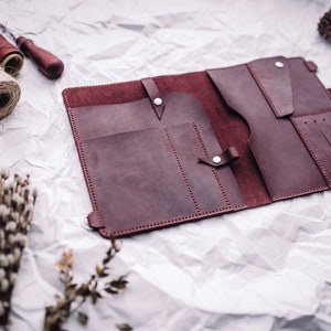 May include: A burgundy leather wallet with multiple pockets and compartments, open and lying flat on a white crumpled paper background.