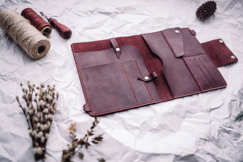 May include: A burgundy leather wallet with multiple pockets and compartments, open and lying flat on a white crumpled paper background.