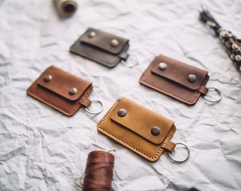 May include: Four leather keychains with a small pocket on the front. The keychains are in different shades of brown and gray. The keychains are on a white crumpled paper background.