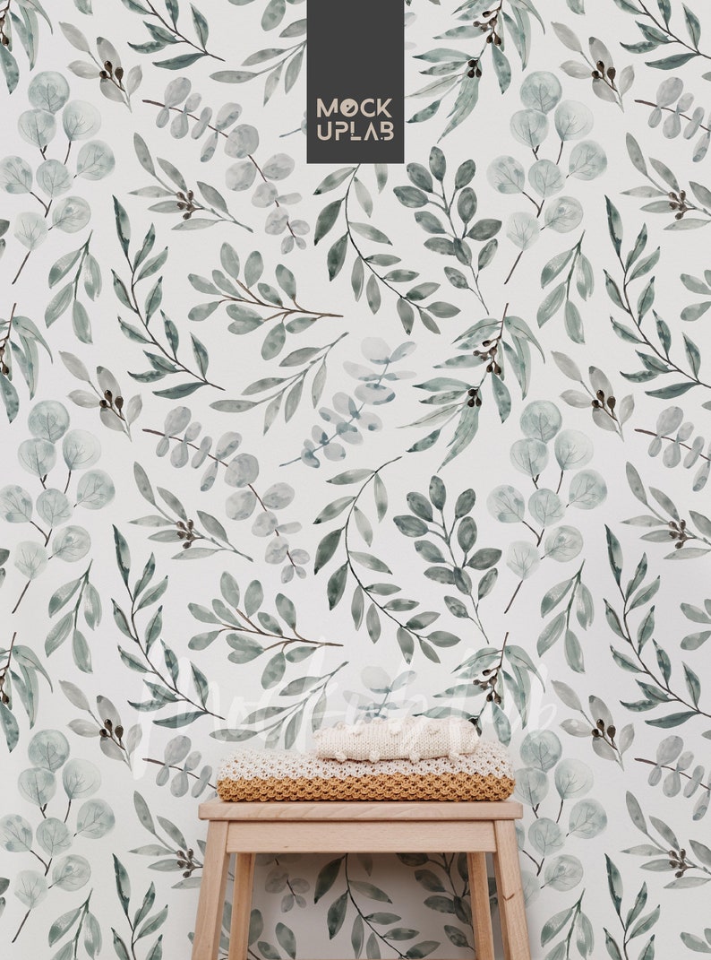 May include: A white wall with a green watercolor leaf pattern. A small wooden stool with a knitted blanket on top sits in front of the wall.