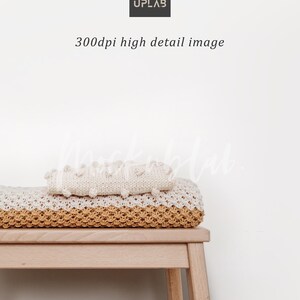 May include: A white knitted blanket with a textured surface sits on top of a yellow knitted blanket. Both blankets are folded and placed on a wooden stool against a white wall.