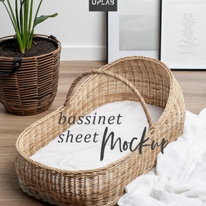 K&ouml;nnte beinhalten: Ein Weidenkorb-Babybett mit einem wei&szlig;en Laken im Inneren. Das Babybett steht auf einem Holzboden mit einer wei&szlig;en Decke daneben. Der Text "bassinet sheet Mockup" ist auf das Laken geschrieben.