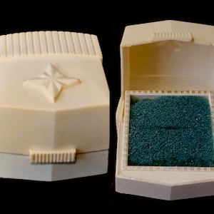 May include: Two vintage ivory jewellery boxes. One is closed, with a starburst design on the lid. The other is open, revealing a teal interior. The boxes are rectangular with a hinged lid.