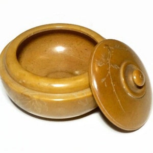 May include: A small, round container with a lid, in a warm, light brown hue. The lid is slightly ajar, revealing the interior. The lid features a carved floral design. The container is likely made of wood or a similar material.