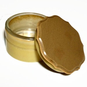 Peut inclure: Un poudrier vintage avec une base jaune clair et un dessus en verre transparent. Le couvercle est en forme de bouclier avec une surface texturée brun clair. Le poudrier est ouvert.