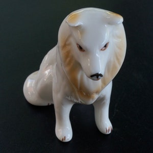 May include: A ceramic dog figurine in a seated pose. The dog is mainly white with tan and brown detailing on its head and paws. It features brown eyes and a black nose. This figurine is likely a decorative piece.