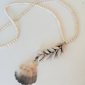 Op de afbeelding: Een witte kralenketting met een grote schelp hanger en een tros kleinere schelpen die eronder hangen.
