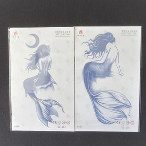 May include: Two temporary tattoo sheets with blue mermaid designs. The left sheet features a mermaid with a crescent moon above her. The right sheet features a mermaid sitting on a rock. Both sheets have the text "GZ-212" and "GZ-216" printed on them.