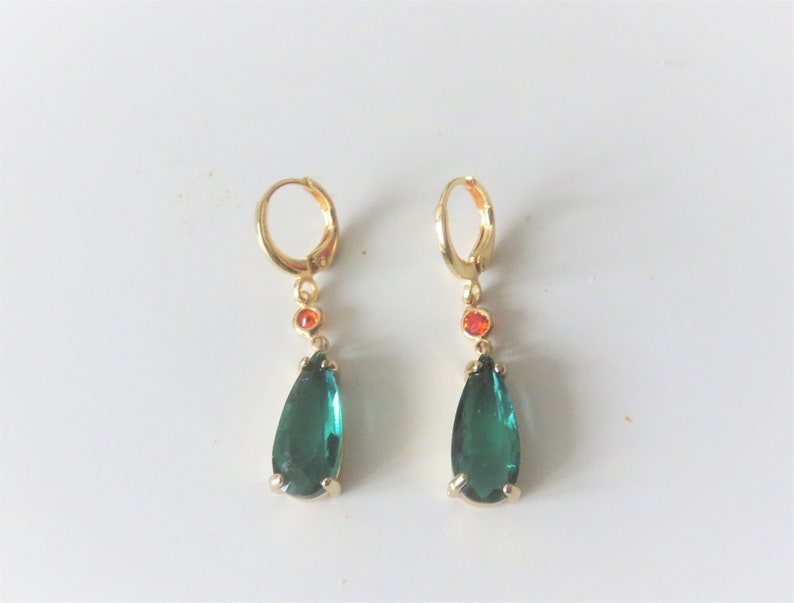 May include: A pair of gold hoop earrings with a teardrop-shaped green gemstone and a small orange gemstone hanging from each hoop.