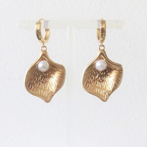 May include: A pair of gold hoop earrings with a single pearl dangling from each hoop. The pearl is nestled inside a gold petal-shaped pendant.