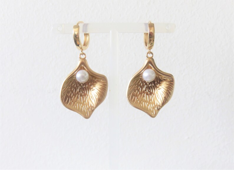 May include: A pair of gold hoop earrings with a single pearl dangling from each hoop. The pearl is nestled inside a gold petal-shaped pendant.