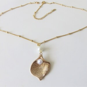 May include: A gold chain necklace with a delicate gold leaf pendant featuring a white pearl in the center. The chain is made of small gold beads.