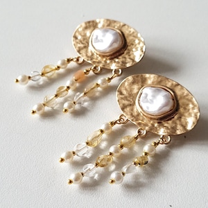 May include: Gold tone earrings with a large oval shaped pendant featuring a white baroque pearl. The pendant is surrounded by a textured gold tone metal. The earrings have multiple strands of white pearls, clear crystals, and gold tone beads hanging from the bottom.