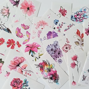 Puede incluir: Surtido de tatuajes temporales con diseños florales y de mariposas en colores rosa, morado y rojo. Los tatuajes están impresos en papel blanco y están listos para ser aplicados a la piel.