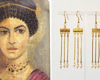 May include: A pair of gold-toned dangle earrings featuring a rectangular textured top bar with three thin hanging chains, each ending in a small white bead. The earrings are displayed on a clear acrylic stand next to a portrait of a person wearing similar jewelry.