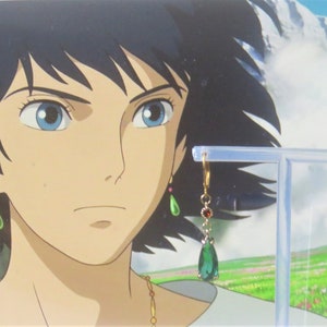 May include: A pair of gold dangle earrings with teardrop-shaped green gemstones and red accents. The earrings are on a display stand against a background of a cartoon character.