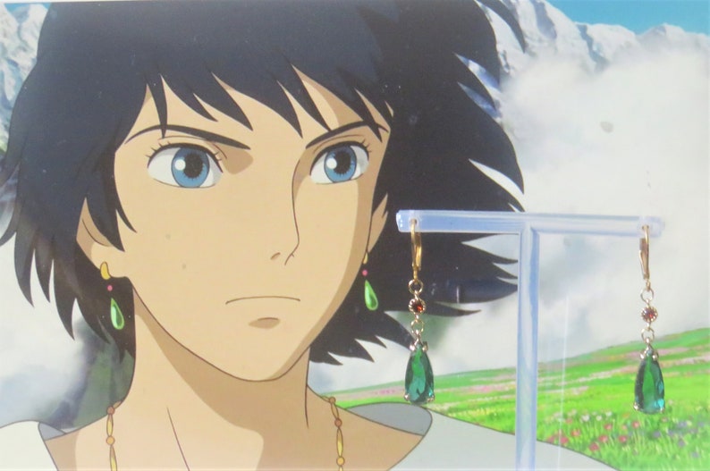 May include: A pair of gold dangle earrings with teardrop-shaped green gemstones and red accents. The earrings are on a display stand against a background of a cartoon character.