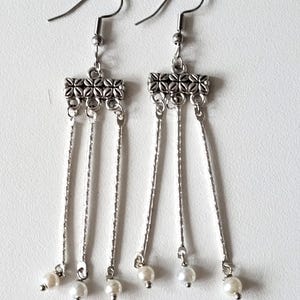 May include: Silver dangle earrings with three chains and a pearl at the end of each chain. The top of the earring has a silver bar with a decorative design.