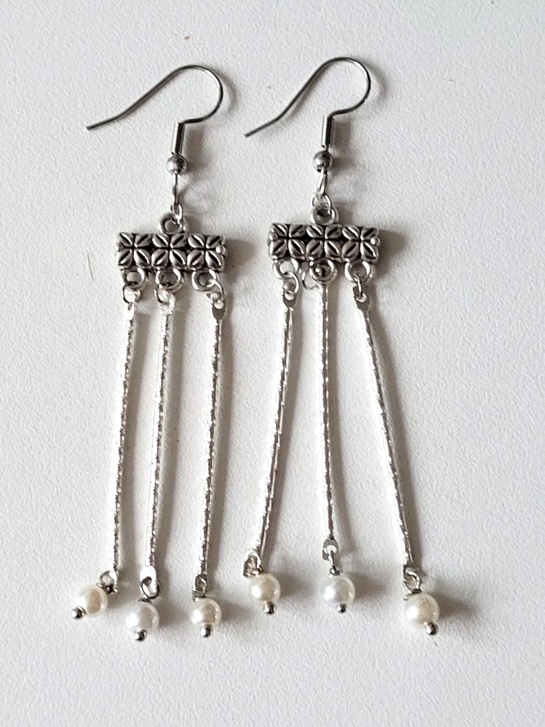 May include: Silver dangle earrings with three chains and a pearl at the end of each chain. The top of the earring has a silver bar with a decorative design.