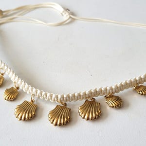 May include: Cream-colored macrame necklace featuring multiple gold-colored seashell charms. The necklace is adjustable with a sliding knot closure. The seashells are detailed with textured ridges, adding a touch of coastal charm to the jewelry.