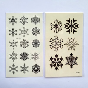 Peut inclure: Deux feuilles de tatouages temporaires avec des motifs de flocons de neige noirs. La feuille de gauche a 18 flocons de neige et le texte "HC-181". La feuille de droite a 6 flocons de neige et le texte "H-083".