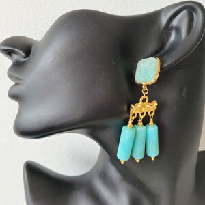 May include: Gold dangle earrings with a turquoise blue square stone and three turquoise blue cylinder beads.
