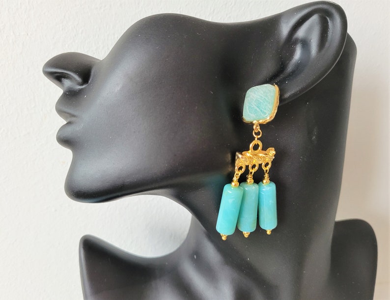 May include: Gold dangle earrings with a turquoise blue square stone and three turquoise blue cylinder beads.