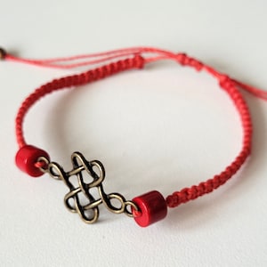 Peut inclure: Bracelet en cordon rouge avec un charme en forme de nœud doré et deux perles rouges.