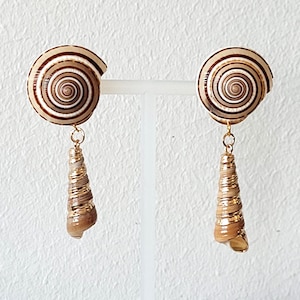 Peut inclure: Une paire de boucles d'oreilles dorées avec une coquille de mer brune et blanche à motif spiralé et une plus petite coquille de mer brune et dorée qui pend en dessous.