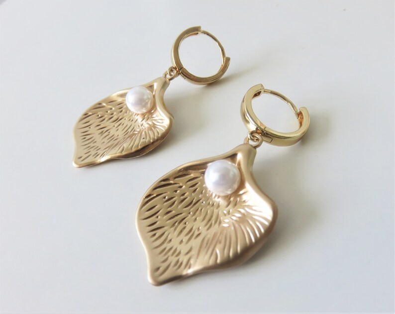 May include: Gold hoop earrings with a textured leaf-shaped pendant and a white pearl accent.