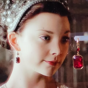 Pode incluir: Uma mulher usando uma faixa de cabeça prateada com um grande brinco de pedra vermelha. O brinco está pendurado em uma corrente de ouro.