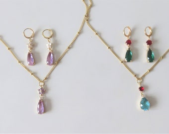 May include: Two gold-toned necklaces and matching earring sets. One set features amethyst-colored teardrop pendants and earrings. The other set has green and red pendants and earrings, and a blue pendant necklace.