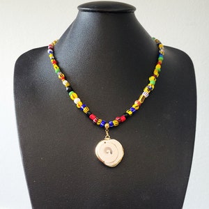 Puede incluir: Un collar de cuentas de colores con un colgante de concha blanca. Las cuentas son de color rojo, amarillo, verde, azul y negro.