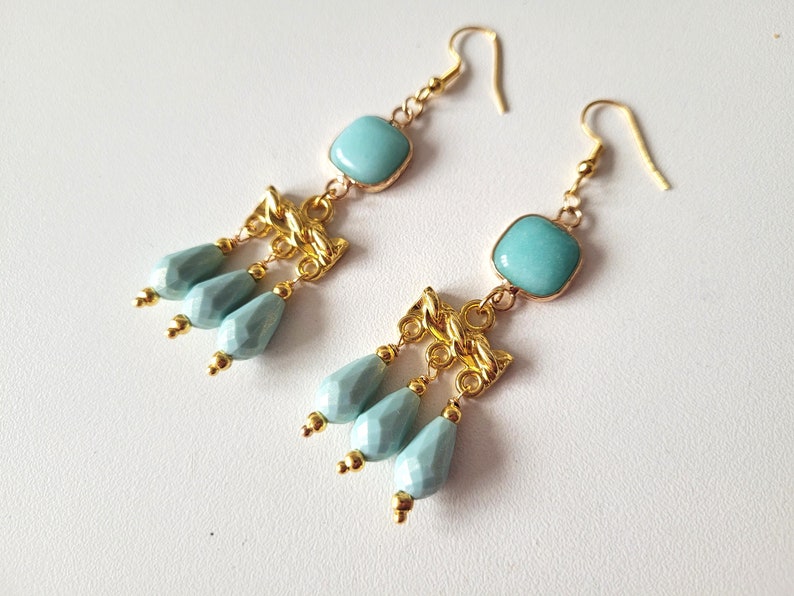 K&ouml;nnte beinhalten: Goldene Ohrh&auml;nger mit einem quadratischen, hellblauen Edelstein und hellblauen, tropfenf&ouml;rmigen Perlen.