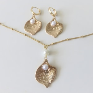 May include: A gold necklace and earrings set with a delicate chain and a pendant shaped like a flower petal with a pearl in the center.