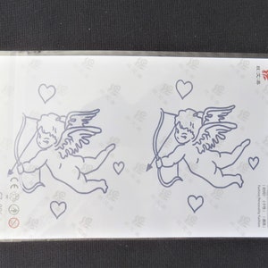 Puede incluir: Dos tatuajes temporales con un cupido con alas y una flecha, rodeado de corazones. Los tatuajes están sobre un fondo blanco.