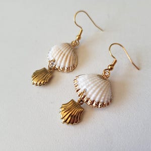 Peut inclure: Boucles d'oreilles coquillage avec crochets et détails dorés. Chaque boucle d'oreille présente un coquillage blanc avec une bordure dorée et un petit pendentif en forme de coquillage doré. Les boucles d'oreilles sont présentées sur une surface blanche.