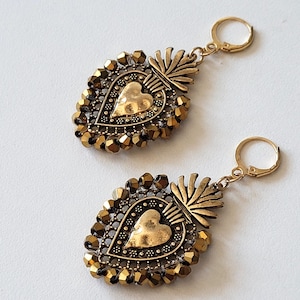 Peut inclure: Une paire de boucles d'oreilles dorées avec un pendentif central en forme de cœur à la texture martelée. Le cœur est entouré d'une bordure de petites perles métalliques à facettes et surmonté d'une crête en éventail. Chaque boucle a un fermoir.