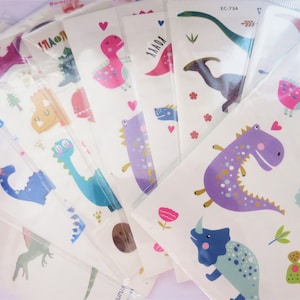 May include: A set of colourful dinosaur temporary tattoos with a variety of designs, including a pink dinosaur with a heart, a purple dinosaur with gold accents, and a blue dinosaur with a rainbow. The tattoos are packaged in clear plastic bags and have the text "Happy Birthday" and "EC-734" and "EC-722" printed on them.