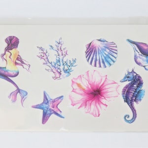 May include: A sheet of temporary tattoos featuring a mermaid, a starfish, a seashell, coral, a hibiscus flower, and a seahorse. The tattoos are in shades of blue, purple, pink, and yellow.