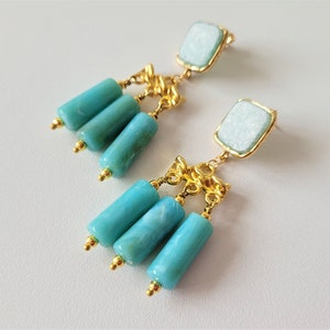 May include: A pair of gold dangle earrings with turquoise blue and white stone accents. The earrings feature a square-shaped stone at the top and three cylindrical stones hanging below.
