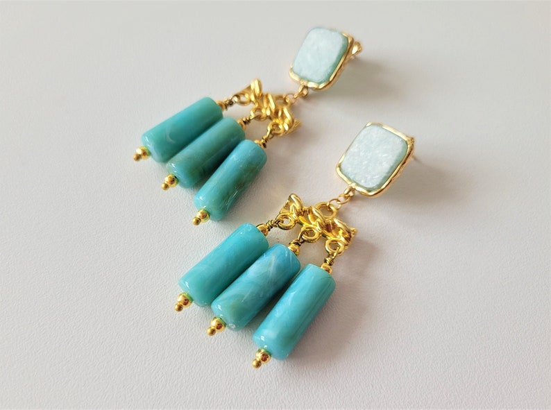 May include: A pair of gold dangle earrings with turquoise blue and white stone accents. The earrings feature a square-shaped stone at the top and three cylindrical stones hanging below.