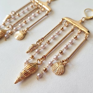 Può includere: Un paio di orecchini color oro a tema conchiglia. Ogni orecchino presenta una barra superiore con aste pendenti, adornate con piccole perle rosa e ciondoli a forma di conchiglia. Gli orecchini hanno una chiusura a leva.