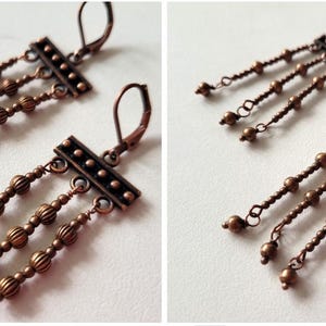 May include: A pair of copper-toned dangle earrings featuring a horizontal rectangular bar with a dotted pattern. Three vertical chains hang from the bar, each adorned with small, round, ribbed beads of varying sizes, finished with lever-back closures.