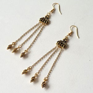 Peut inclure: Une paire de boucles d'oreilles pendantes dorées. Chaque boucle d'oreille présente un dessus filigrané orné, trois délicats brins de chaîne et des breloques en forme de goutte. Les boucles d'oreilles sont présentées sur une surface blanche.